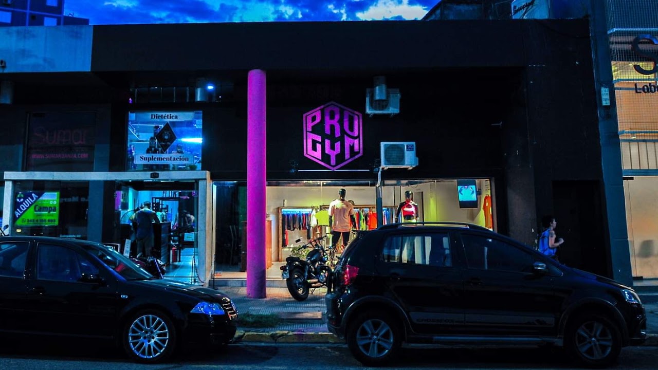 Fachada nocturna de Pro Gym Paraná con cartel de neón rosa y ventanales panorámicos hacia la sala de pesas